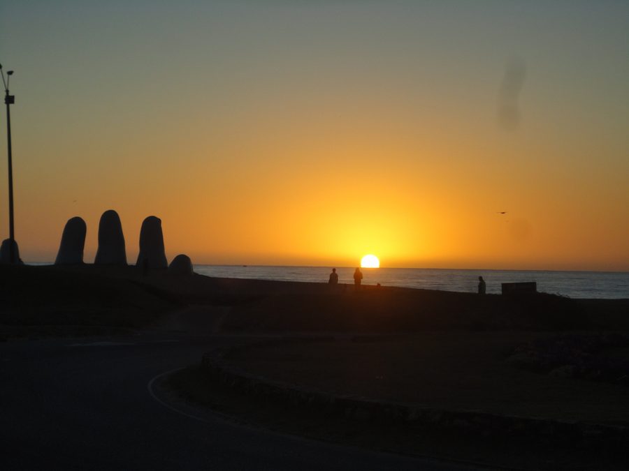El sol saliendo del océano sobre la Playa Brava de Punta del Este, con la silueta de los dedos de La Mano recortada contra la luz