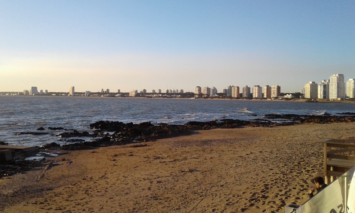 La Playa Mansa de Punta del Este en una tarde de julio: arena vacía, rocas oscuras, agua quieta y las torres del balneario al fondo bajo un cielo despejado