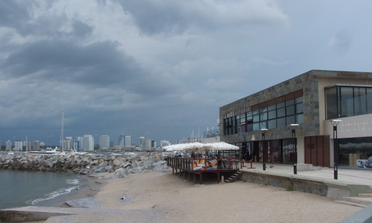 Un restaurante sobre la arena junto al puerto de Punta del Este, con sombrillas blancas en la terraza y las torres del balneario al fondo