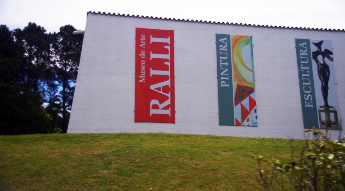 El muro blanco del Museo Ralli de Punta del Este sobre el césped, con los carteles de pintura y escultura y los pinos del parque al costado