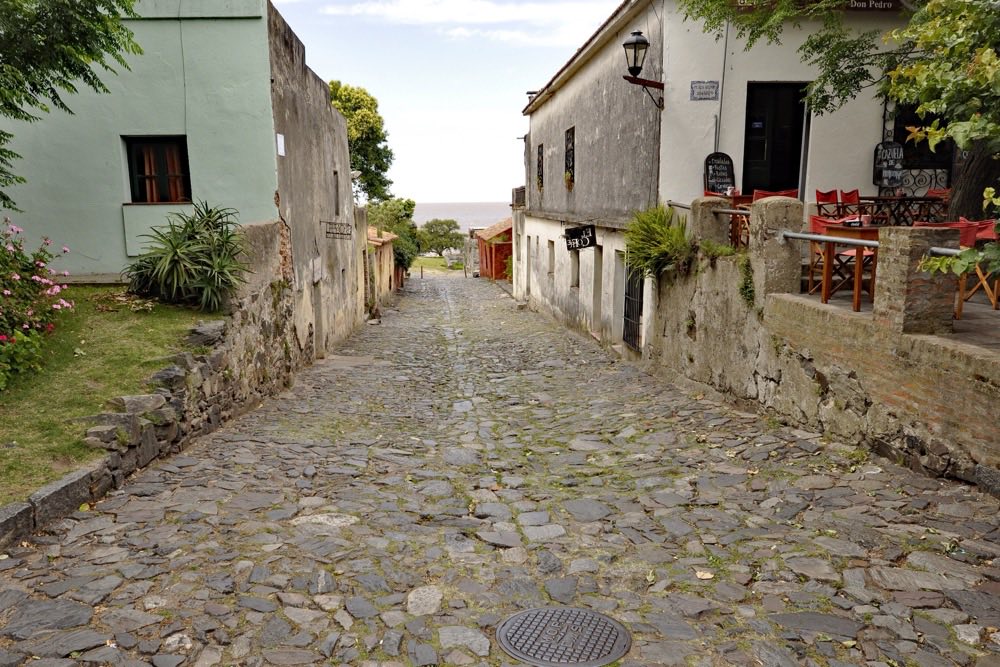 Calle de piedra del barrio histórico de Colonia del Sacramento, con casas coloniales bajas y el río al final de la cuesta